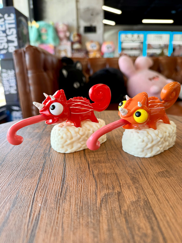 Two cartoonish chameleon figurines, Sushi Gogo Chameleons, perched on sushi rice mounds, with curled tongues and horn-like protrusions, displayed on a wood-grain tabletop.