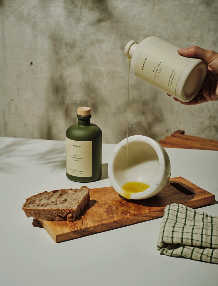 Olive oil being poured from bottle into a glass cup, with bread and a cutting board