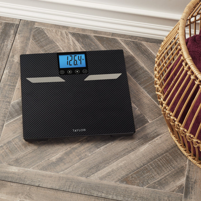 Top-down view of Taylor carbon fiber digital scale on tile floor with a wicker basket nearby