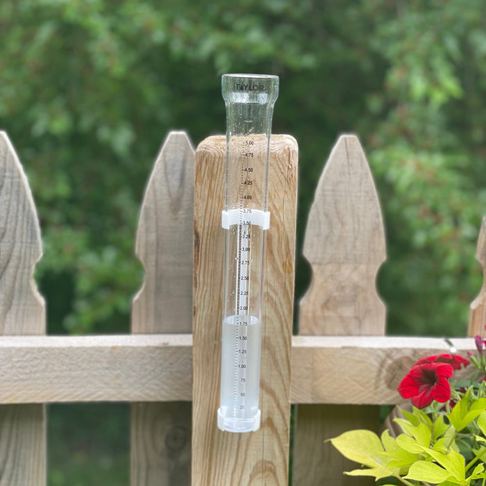 ClearVu rain gauge attached to a wooden fence post in a garden with greenery in the background and a red flower nearby