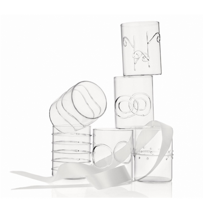 Six clear borosilicate glass tumblers from the Deco Clear Water Glass Set of 6 by Ichendorf Milano, each featuring distinctive etched or molded designs, are stacked atop one another. A white ribbon winds through and around the glasses, elegantly draping across their surfaces. The background is a clean white.