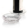 Two round Mille Nuits Wine Coasters by Baccarat, featuring a ridged design, rest on a white surface—one empty in the foreground and the other holding a dark crystal bottle in the background.