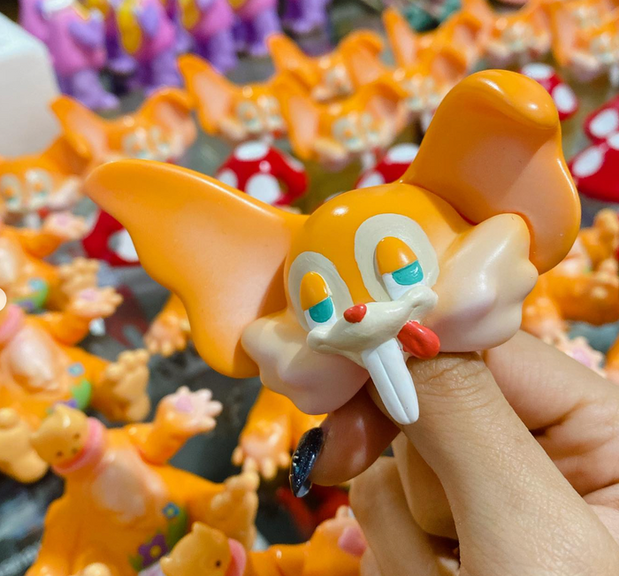 Person holding a Mushroom by Vivian Wang toy head; features buck teeth and floppy ears, with a display of similar figures in the background.